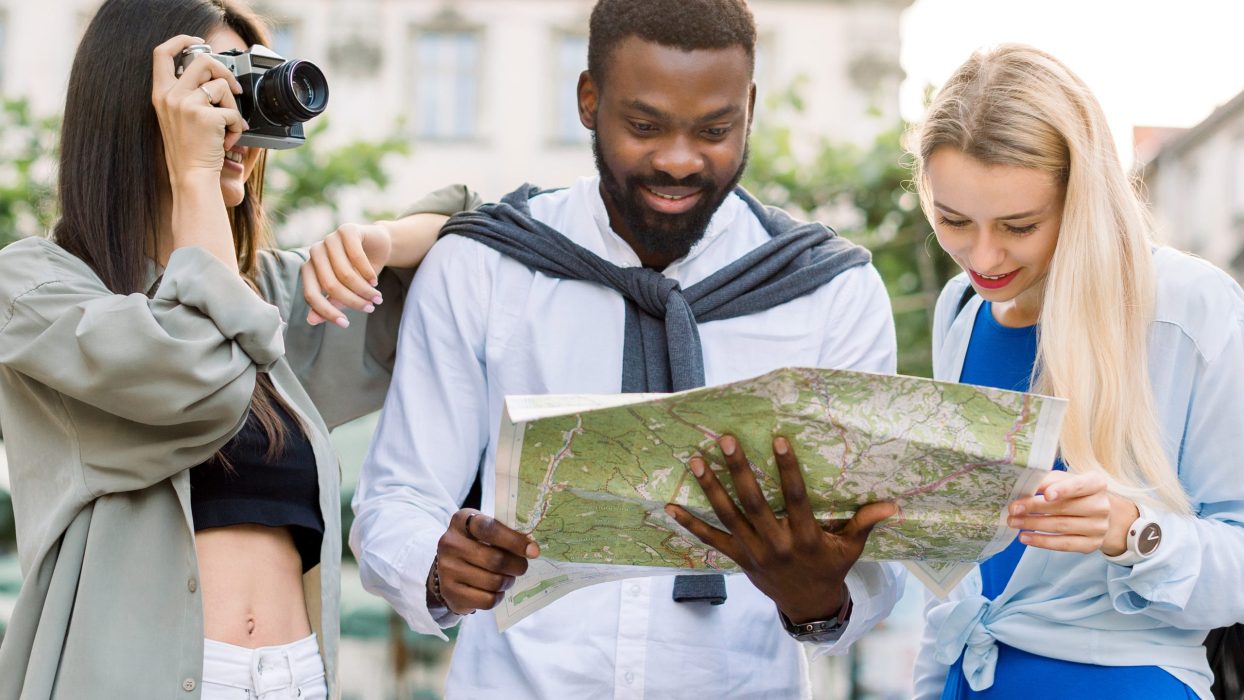 Happy group of multiethnic tourists enjoying sightseeing together in ancient European city. African man and Caucasian blond girl looking at map, while Caucasian brunette girl makes photos on camera.