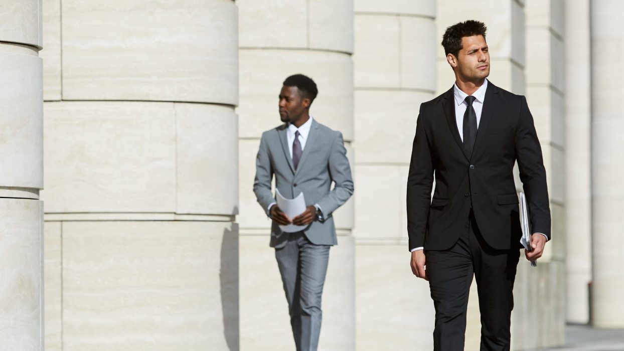 Serious confident businessman in black suit walking along the street in the city with other man in the background