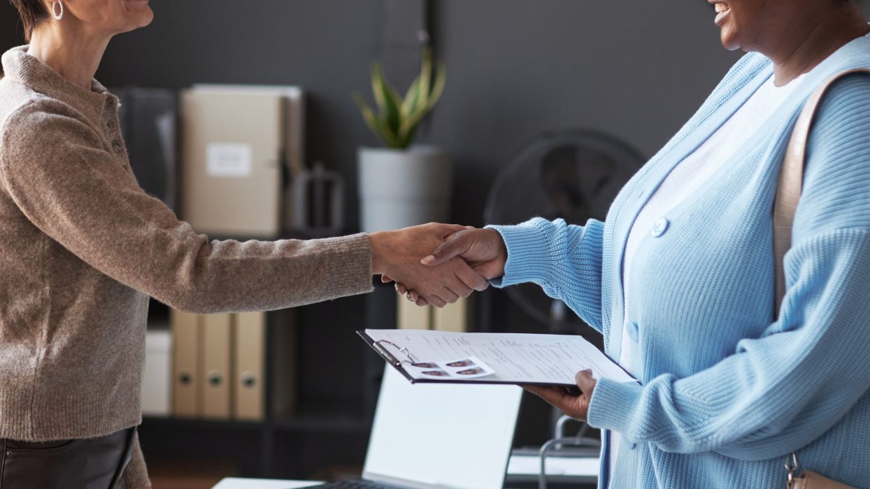 Happy mature manager congratulating young woman on approving visa application while shaking her hand after stamping documents