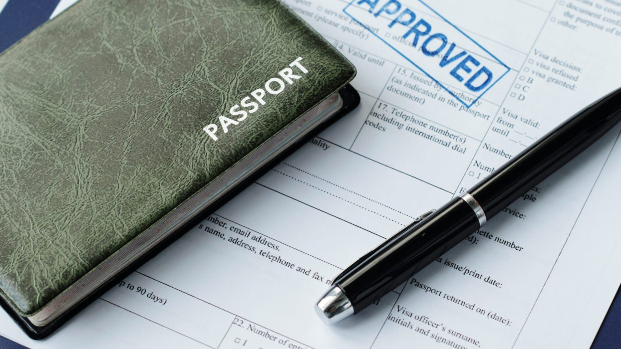 High angle close-up of passport in leather cover, travel visa application paper with approved stamp on it and pen on table