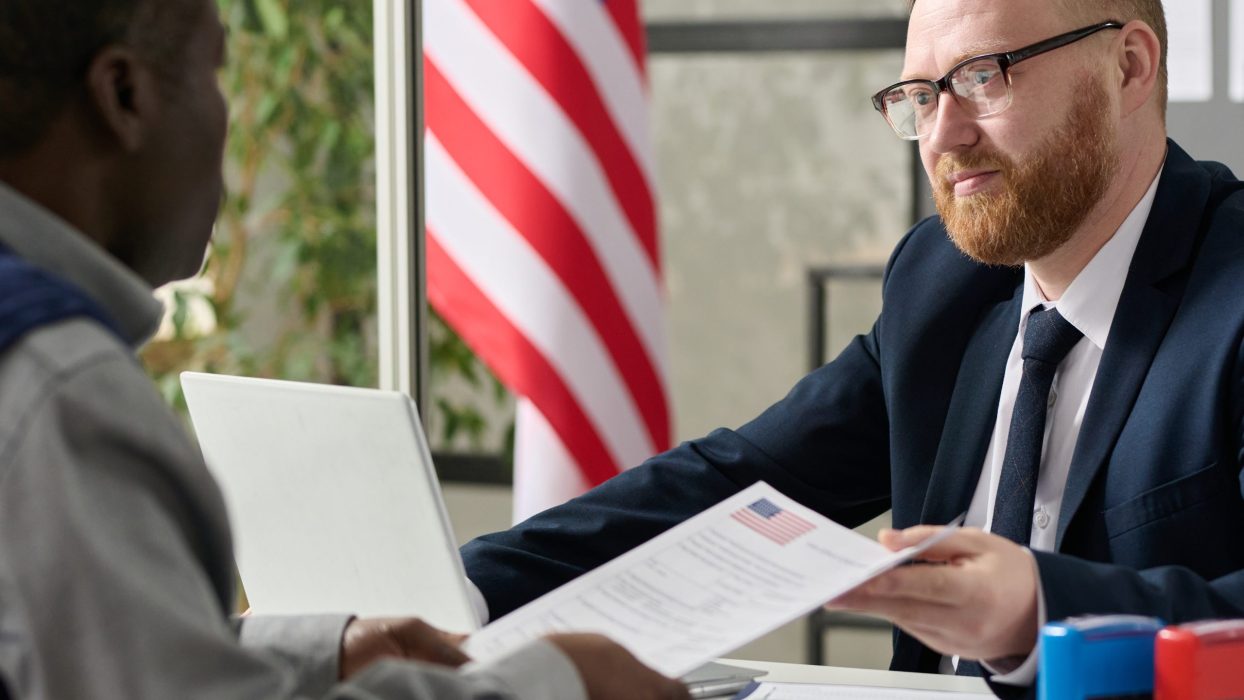 Portrait of bearded male worker handing visa application to black man in US immigration office